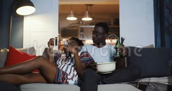 快乐的年轻非洲积极的朋友夫妇在家里沙发上看电视吃零食和笑慢动作视频的预览图