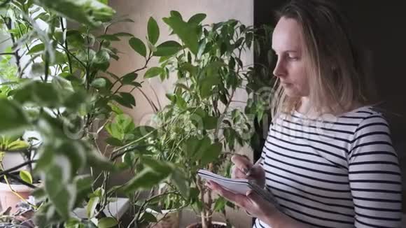 穿背心的漂亮女孩检查她家的植物年轻女性在家里清点植物和进入视频的预览图