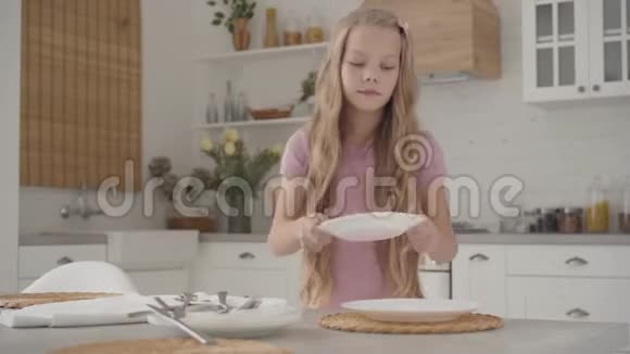 年轻的金发白种人女孩穿着粉红色的裙子端着桌子微笑着勤奋的青少年在家里做家务视频的预览图