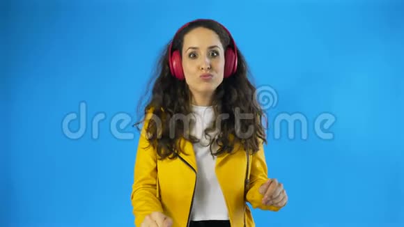 穿着黄色夹克的滑稽可爱的女人正在蓝色背景的工作室里用耳机听音乐跳舞视频的预览图