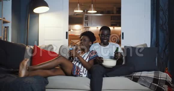 快乐的年轻非洲千禧浪漫情侣在家沙发上看电影吃零食说话慢动作视频的预览图