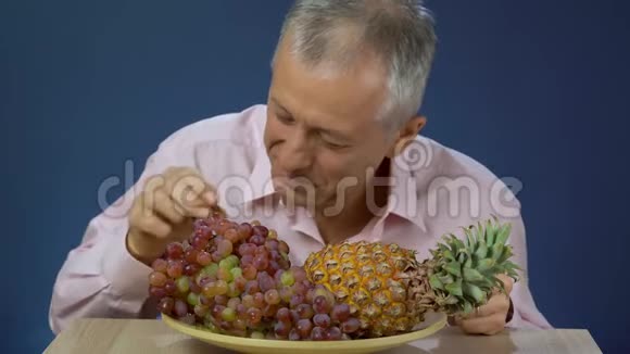 一个穿着衬衫的中年男人高兴地吃着放在菠萝旁边的大盘子上的葡萄微笑着给了一个视频的预览图