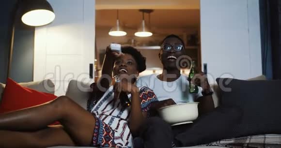 快乐年轻积极的黑人朋友男人和女人看电视在家里用遥控器拿零食和饮料慢动作视频的预览图