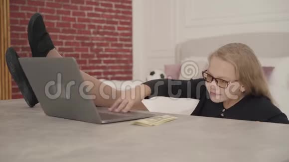 快乐的白人少女坐在她的房间里拿着笔记本电脑和零花钱双腿自信的女学生视频的预览图