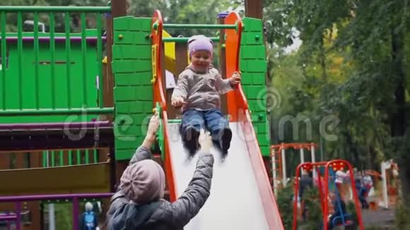 可爱的白种人幼儿男孩在城市公园的操场上玩着滑下潮湿的金属滑梯视频的预览图