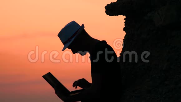 迷人的年轻人带着笔记本电脑坐在海边橙色日落的岩石上快乐度假时光概念男性视频的预览图