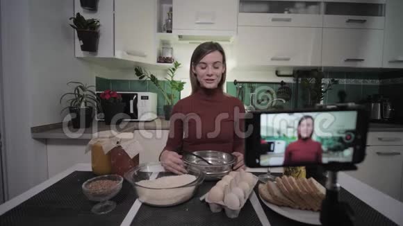 一位美丽的女性博主坐在餐桌上厨房里摆着杂货为她的用户写智能手机视频食谱视频的预览图