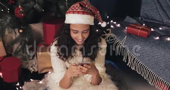 戴着圣诞帽的美女坐在附近地板上用智能手机打字问候朋友视频的预览图