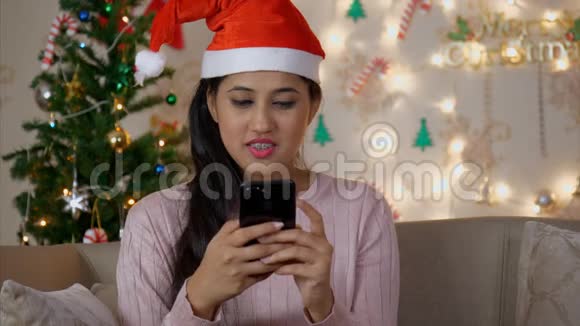 靠近一个年轻快乐的女人在她的手机上打字聊天背景是圣诞树视频的预览图