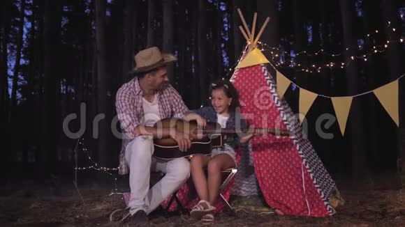 在森林里露营快乐的父亲和可爱的女儿在wigwam附近唱歌弹吉他上面装饰着花环视频的预览图