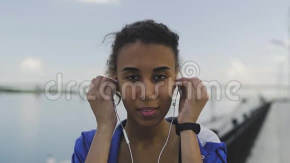 年轻的非裔美国妇女正在看着摄像机戴着耳机为心脏训练提供音乐动力河流视频的预览图