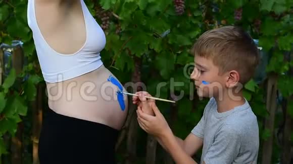 笑的男孩用刷子在他怀孕妈妈宝宝的肚子上画脚印怀孕的概念特写视频的预览图