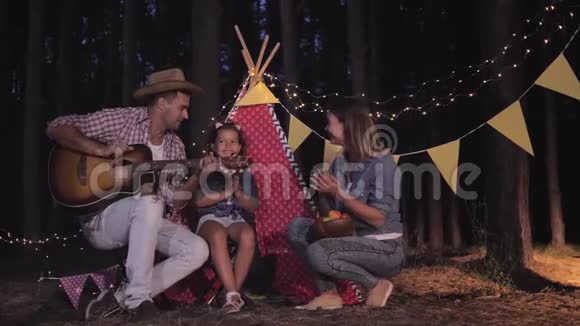 快乐的父亲在森林里为快乐的妻子和可爱的女儿在野餐时弹吉他用火歌唱视频的预览图