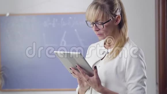 白种人中年妇女站在平板电脑和看设备屏幕的肖像优雅的私人教师视频的预览图