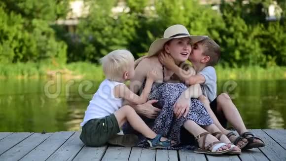两个小男孩在妈妈耳边低语夏日河岸上的码头幸福的家庭视频的预览图