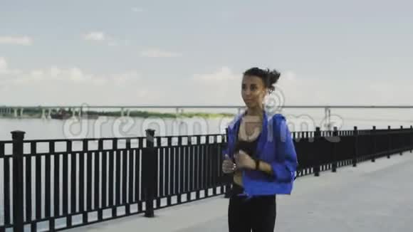 穿着蓝色夹克带耳机的美国黑人女跑步者沿着堤道跑步视频的预览图