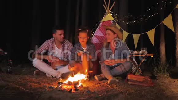 夜晚野餐时可爱的女儿和快乐的父母围着火堆聊天喝果汁煎棉花糖视频的预览图