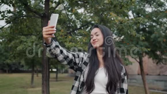 有魅力的亚洲女孩用智能手机相机在公园自拍的肖像视频的预览图