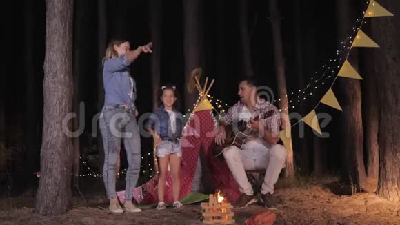 快乐的爸爸为家人弹吉他妈妈和女儿周末在森林里的篝火晚会上尽情跳舞视频的预览图