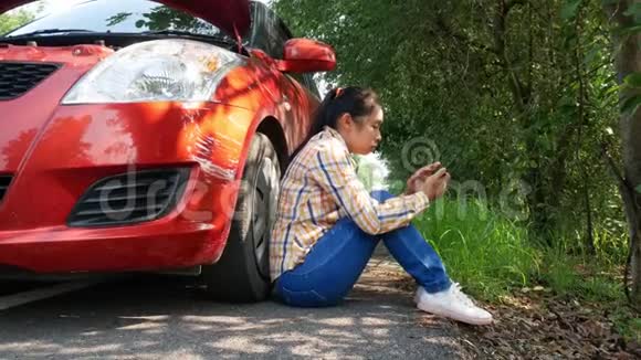 亚洲年轻的女人一边使用智能手机一边坐着等待汽车机械师在路上的断车附近视频的预览图