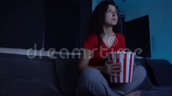 晚上女孩在沙发上看电视上的恐怖电影从桶里吃爆米花害怕躲在一桶水后面视频的预览图