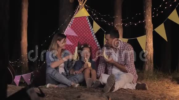 家庭露营快乐的爸爸妈妈和美丽的女儿在森林里的夜晚野餐吃香蕉笑视频的预览图
