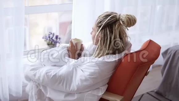 金发女孩长着长长的可怕的眼睛望着窗外舒适的公寓坐在大窗户旁边的女人拿着杯子视频的预览图