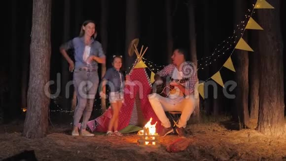 家庭的乐趣快乐的父亲为妈妈弹吉他女儿在夏季周末在篝火旁跳舞视频的预览图