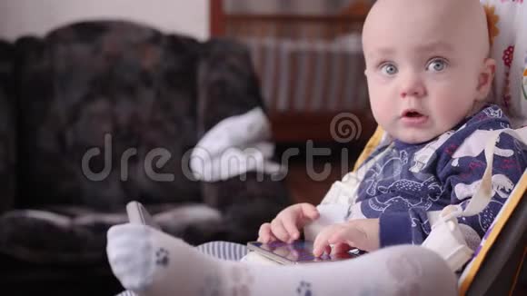 一个光头的小男孩在第一次理发后坐在儿童椅子上玩智能手机慢慢视频的预览图