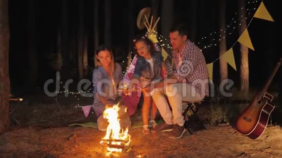 家庭气氛有趣的父母带着他们的小女儿在树桩上烤棉花糖在森林里欢笑一夜视频的预览图