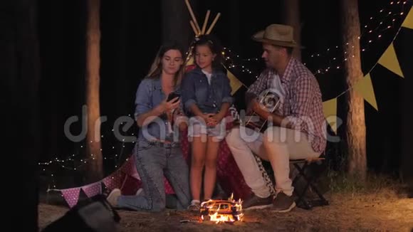家人自拍微笑的爸爸妈妈带着迷人的女儿在森林里篝火旁拍照晚上休息视频的预览图