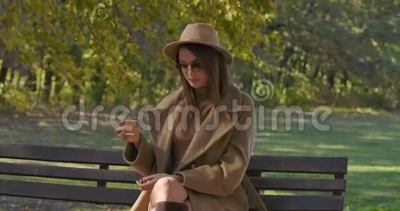 一位优雅的白人妇女戴着棕色帽子和太阳镜坐在长凳上喝茶或咖啡美丽的欧洲人视频的预览图