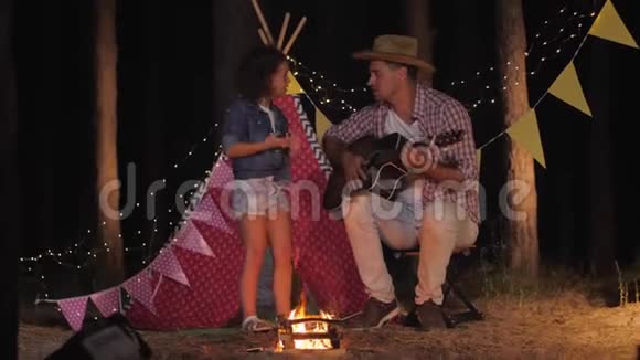 爱的父亲在wigwam的背景下在靠近火堆的树林里露营时为他微笑的女儿弹吉他视频的预览图