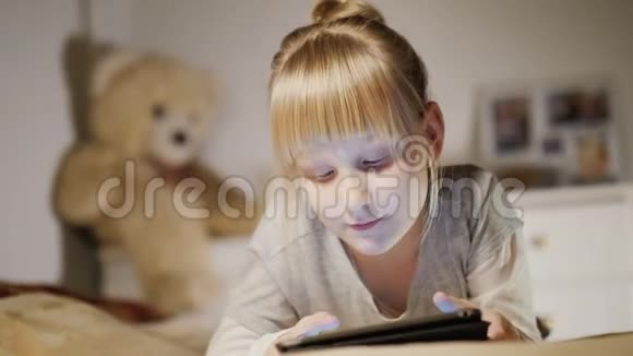 一个6岁的女孩躺在床上躺在房间里的平板电脑上她的玩具从后面可以看到视频的预览图