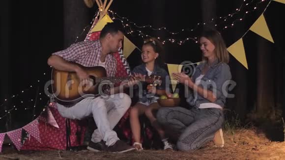 在森林里的家庭带着女孩子的年轻父母在森林里的野餐中玩吉他和吃水果视频的预览图