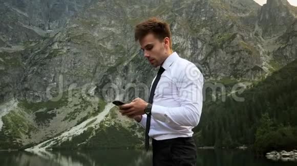 日落时分成功自信的商人在落基山脉的一个湖附近使用一部手机特写照片视频的预览图