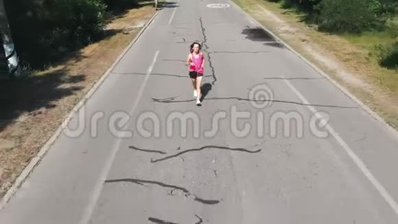 穿着粉红色衬衫的漂亮苗条的女孩正在练习跑步戴着耳机的年轻黑发女子在炎热的夏日在p视频的预览图