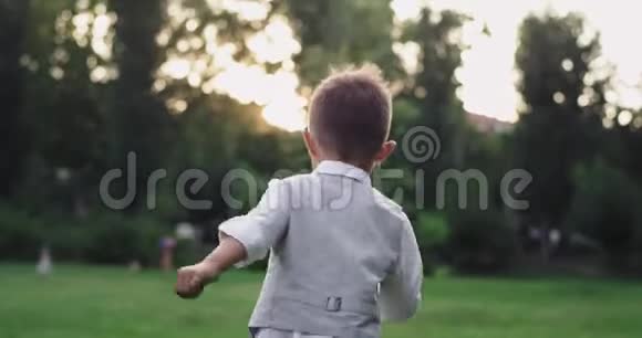 穿着西装跑过公园中央绿草地的小可爱男孩享受着大自然的时光视频的预览图