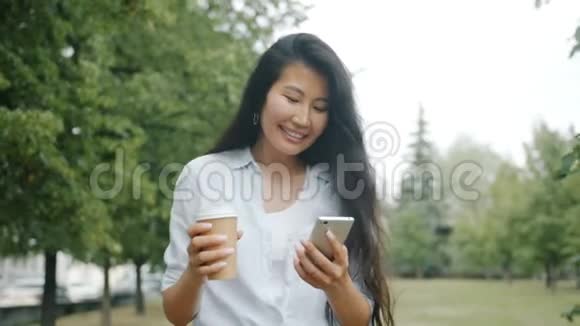 可爱的亚洲学生在公园里散步喝咖啡和智能手机视频的预览图
