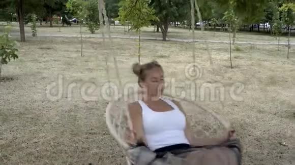 一个性感的金发女郎穿着白色的t恤在一个夏天的晚上在一个城市公园的绳子秋千上摇摆她若有所思地抬起头来视频的预览图