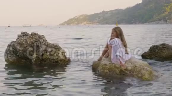 穿着白色长袍的放松的女孩在水中的石头上欣赏海洋景观快乐的少女坐在海边的岩石上视频的预览图