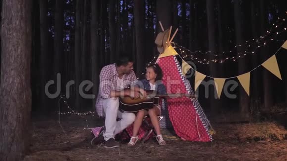 家庭露营在林地爸爸教女儿弹吉他坐在wigwam附近与花环户外视频的预览图