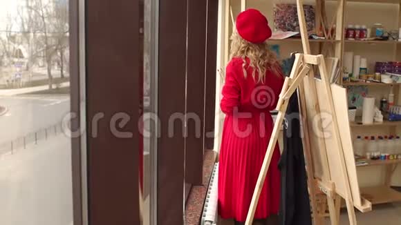 一个穿着红色连衣裙和贝雷帽的女孩站在画室的一个大窗户附近视频的预览图
