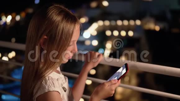 年轻女孩在黑暗阳台上的社交网络中看智能手机在线广播使用手机的少女视频的预览图