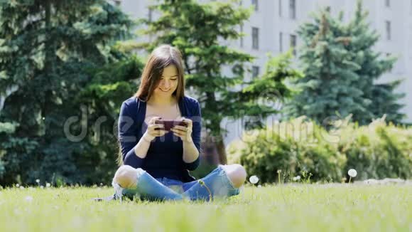 快乐的女人在草地上的城市公园里玩手机游戏视频的预览图