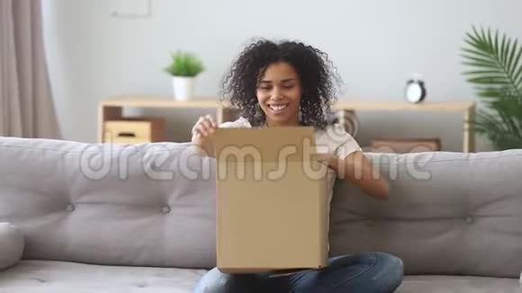 坐在沙发上的快乐混血女人打开收到的包裹视频的预览图