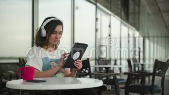 戴着耳机的年轻女子喜欢音乐在咖啡馆里使用平板电脑视频的预览图