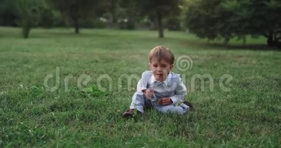在公园里一个穿着休闲服躺在草地上享受零食的愤怒而又风趣的两岁男孩视频的预览图