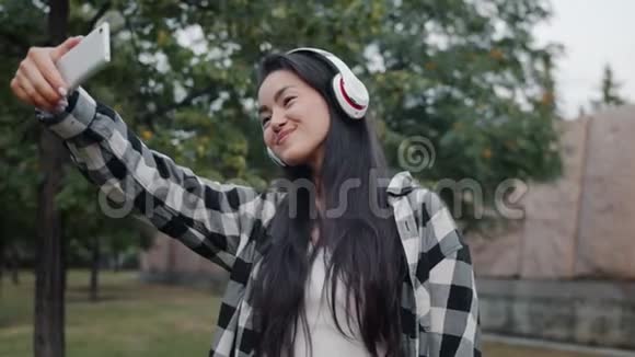 戴着耳机的亚洲女孩在户外用智能手机拍照自拍视频的预览图