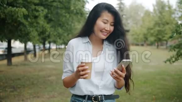 年轻的亚洲女士在公园里用智能手机拿着咖啡视频的预览图